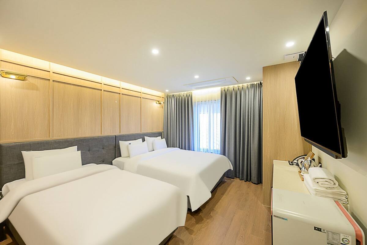 Ein Hotelzimmer mit zwei Betten, einem Flachbildfernseher, einem Schreibtisch mit Kühlschrank und einem Fenster mit Vorhängen.