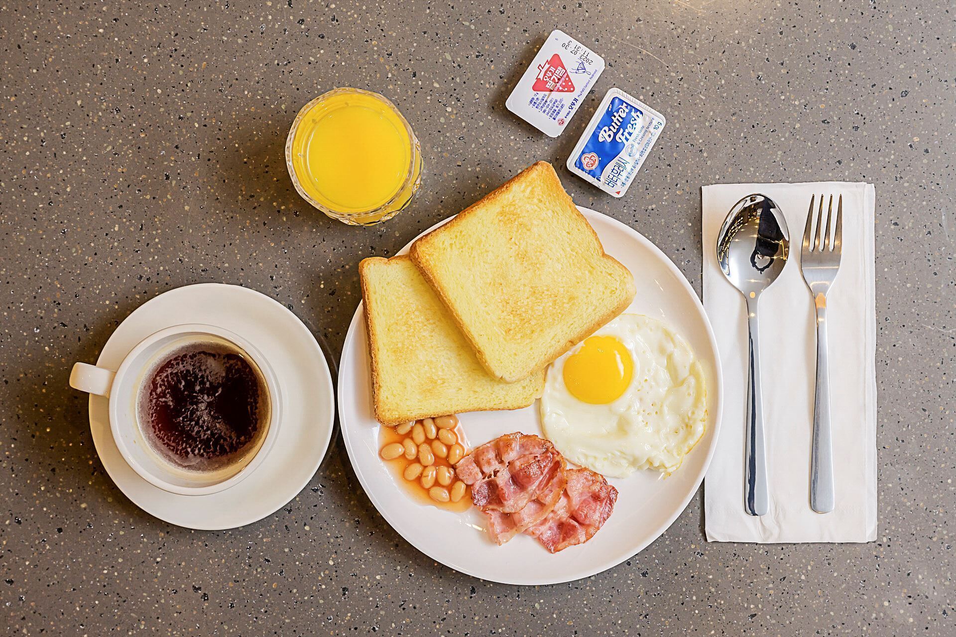 Prato de café da manhã com torradas, ovos, feijão, bacon, uma xícara de café e um copo de suco de laranja em uma bancada texturizada.