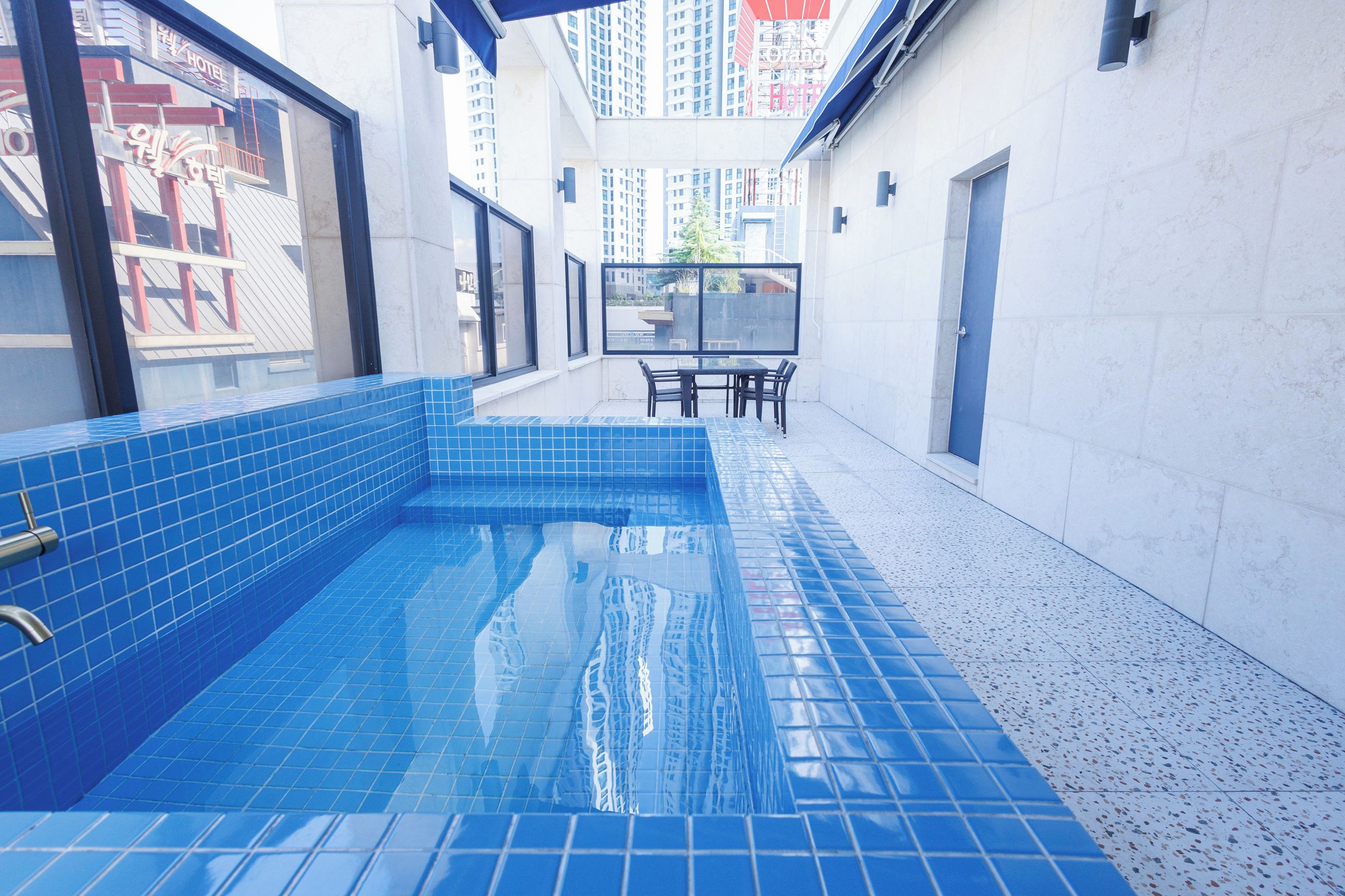 A swimming pool with blue tiles, a table with chairs, and a view of surrounding buildings.