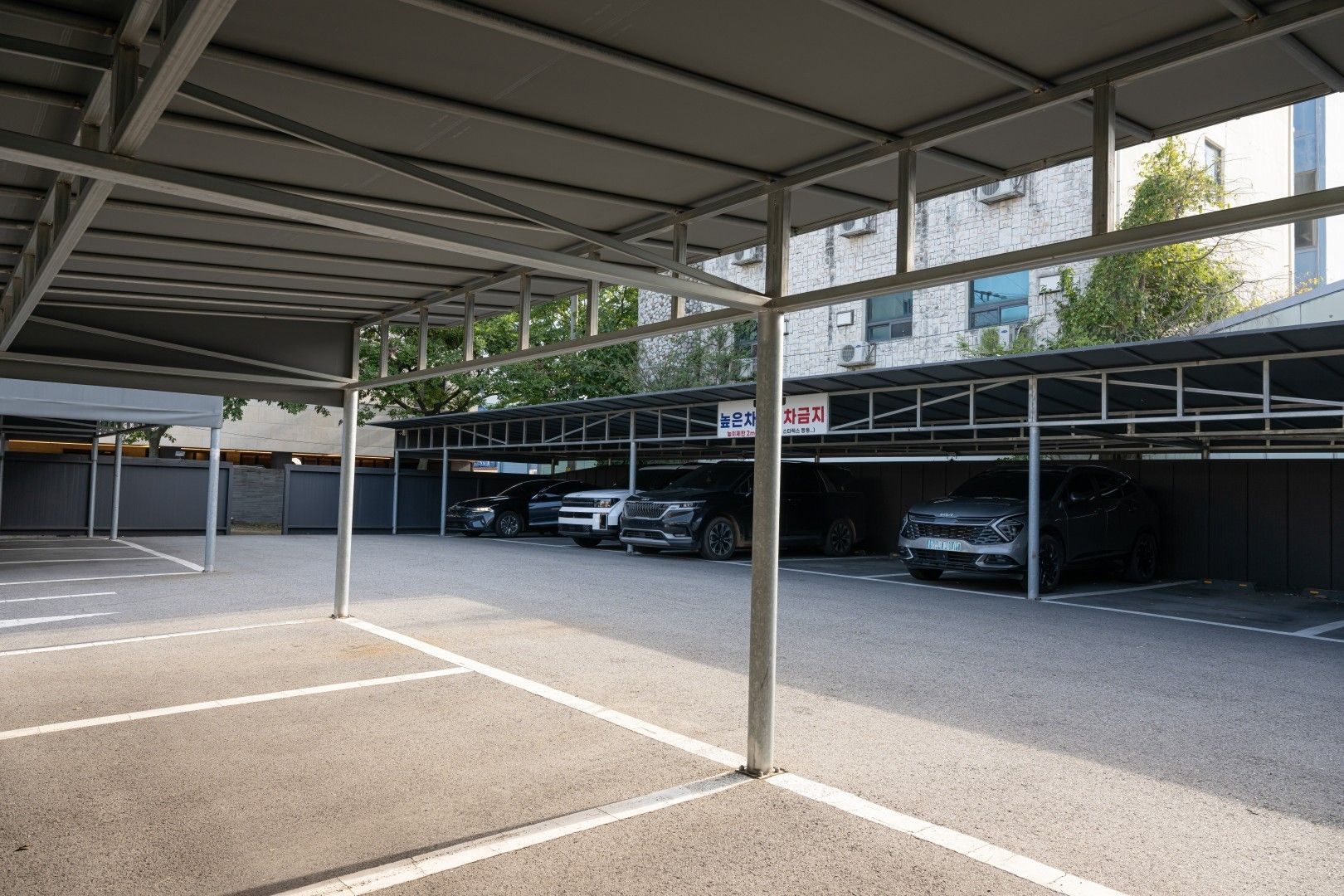 Un espace de stationnement couvert où sont garées plusieurs voitures, des poutres métalliques soutenant le toit, et un bâtiment avec une façade en pierre en arrière-plan.