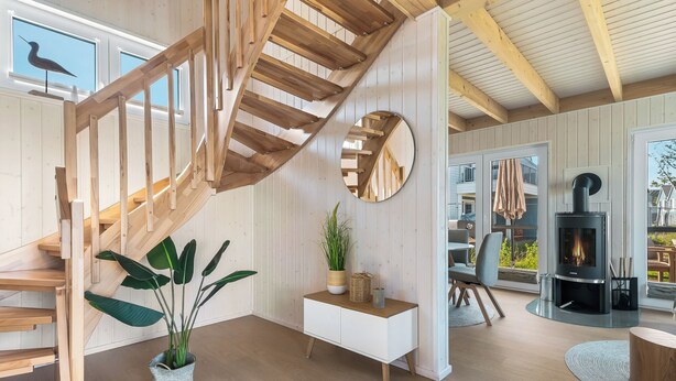Un intérieur moderne avec un escalier en bois, une console blanche agrémentée d’une plante, un miroir rond et une cheminée.