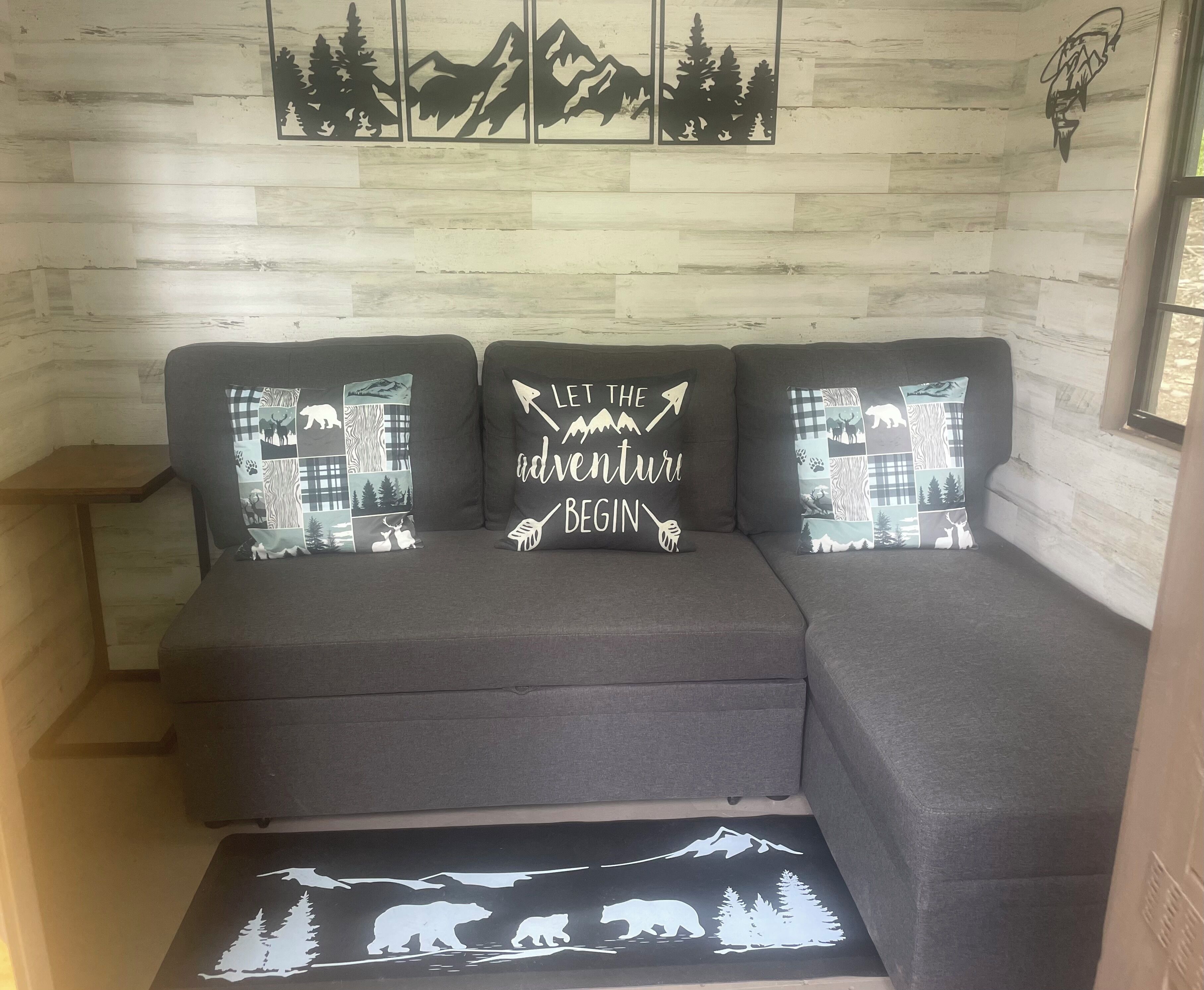 Ein graues Ecksofa mit drei Zierkissen, die Tier- und Naturmotive zeigen, vor einer Holzwand mit einem Berggemälde.