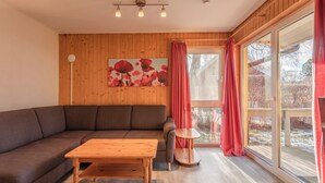 A modern living room with a wooden floor, a brown sofa, a wooden coffee table, a small round table, and a large window with red curtains. - Möwe 35 - Kleines Gemütliches Ferienhaus im Ferienpark Mirow (Mirow)