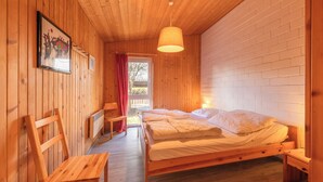 A wooden cabin bedroom with a single bed, a wooden chair, a small table, and a window with a view of the outdoors. - Möwe 35 - Kleines Gemütliches Ferienhaus im Ferienpark Mirow (Mirow)