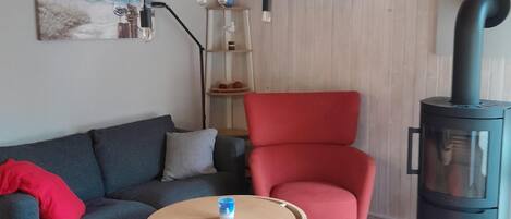 Uma sala de estar moderna com teto de madeira, um sofá vermelho, uma poltrona vermelha, uma mesa de centro redonda de madeira e uma lareira a lenha preta.