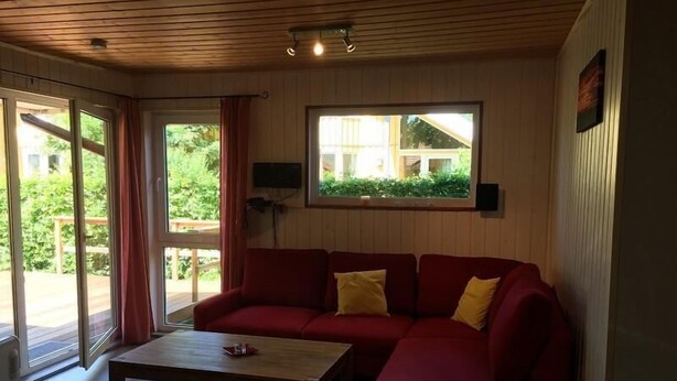 Ein gemütliches Wohnzimmer mit einem roten Sofa, einem Holztisch und einem großen Fenster mit Blick auf Grünflächen.