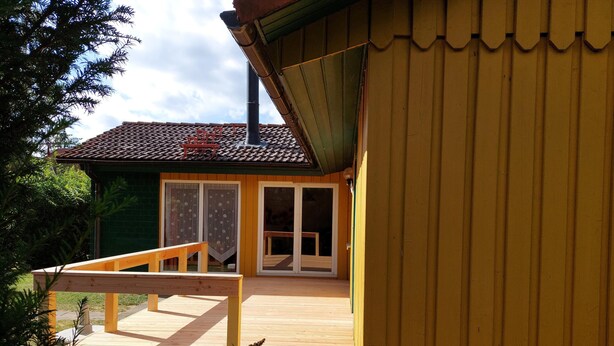 Eine Holzterrasse mit einer Bank, ein grünes Gebäude mit Ziegeldach und eine gelbe Wand.