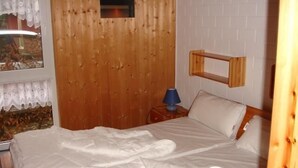 A bedroom with a wooden headboard, a bed with white bedding, a nightstand with a lamp, and a window with a view of trees. - Möwe 151 - Kleines Gemütliches Ferienhaus im Ferienpark Mirow (Mirow)