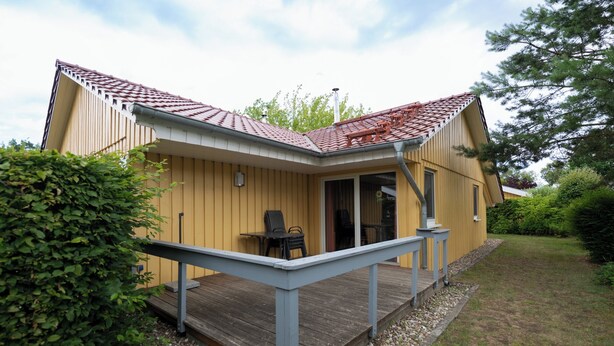 A yellow building with a red-tiled roof, a wooden deck, and a covered outdoor seating area. - Möwe 151 - Kleines Gemütliches Ferienhaus im Ferienpark Mirow (Mirow)