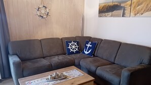 Una sala de estar con un sofá seccional marrón oscuro, dos almohadones decorativos con motivos náuticos y una mesa de centro de madera con periódicos y una bandeja.