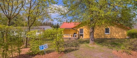 A yellow building with a red roof, surrounded by trees and a paved area with a parking sign.
