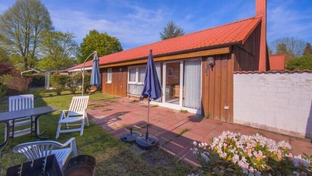 Una casa de una planta con techo de tejas rojas, un patio con mobiliario exterior y un jardín con flores.