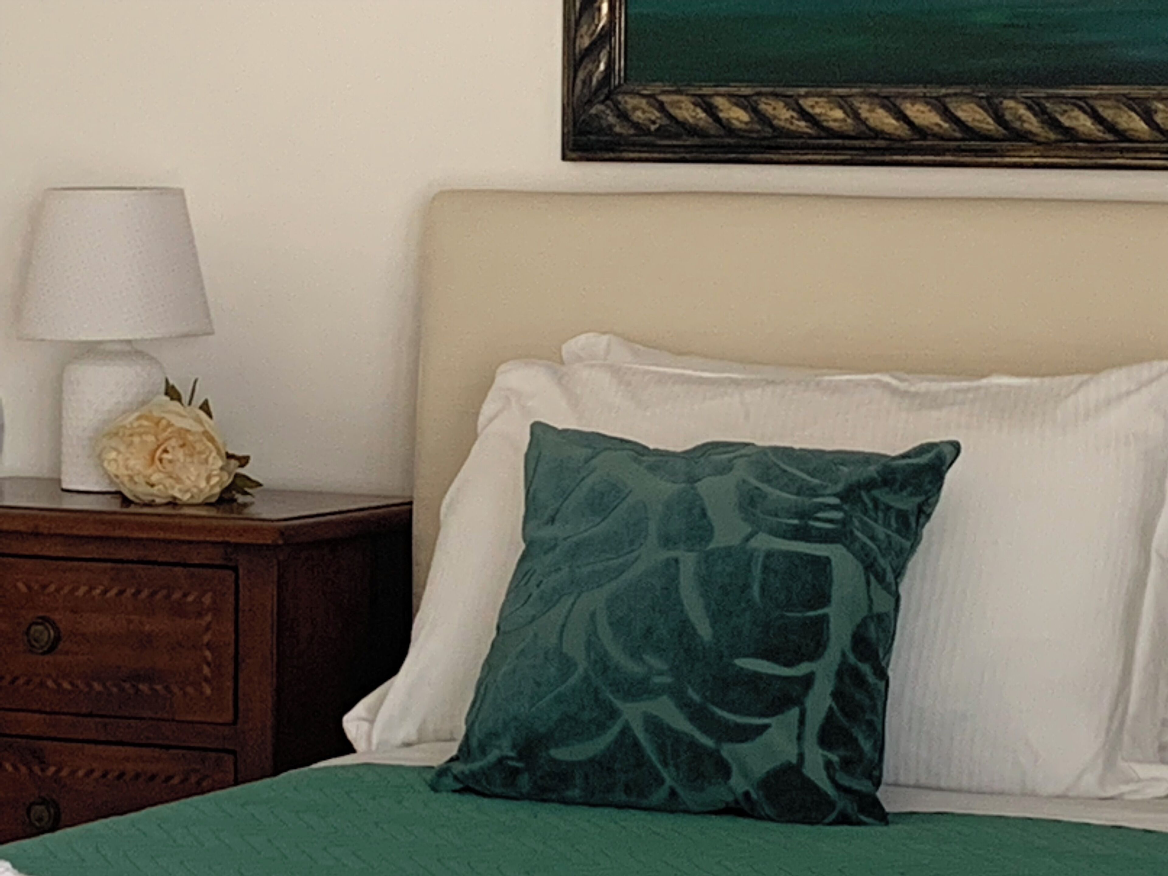 A neatly made bed with a green pillow and a white pillow, a bedside table with a lamp and a flower, and a framed picture on the wall.