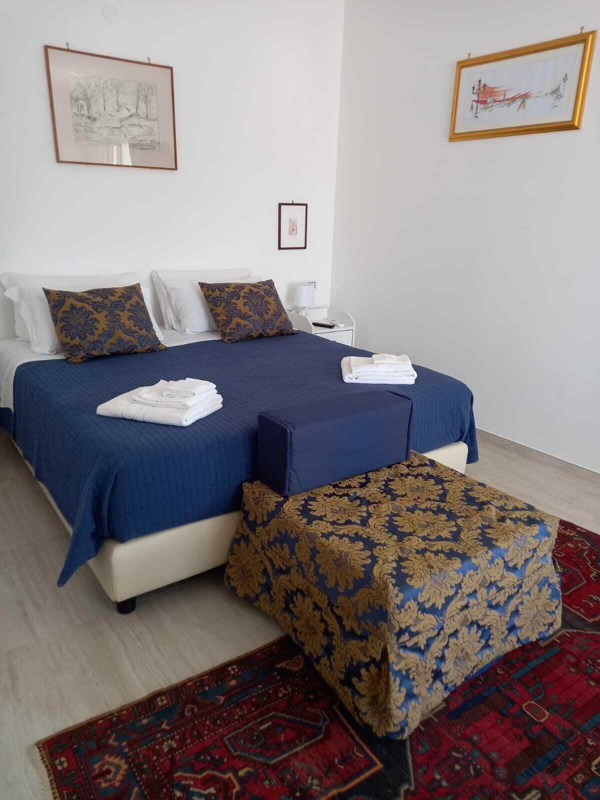 A neatly made bed with blue bedding and two pillows, a bedside table with a lamp, and a red patterned rug on the floor.