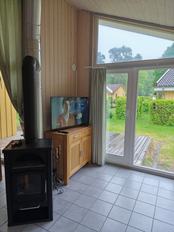 A room with a wood-burning stove, a television, and a view of a garden through a glass door. - Seeschwalbe 94 - Das Skandinavische Familienhaus für 4 mit Sauna und Whirlpool (Mirow)
