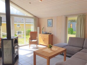 A living room with a sofa, a wooden table, a wicker chair, a cabinet, and a fireplace. - Seeschwalbe 94 - Das Skandinavische Familienhaus für 4 mit Sauna und Whirlpool (Mirow)