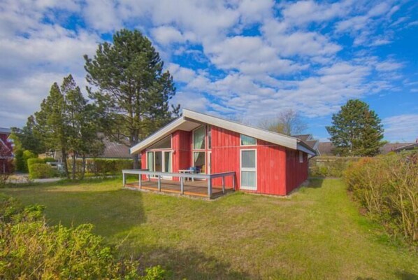 A red cabin with a balcony, surrounded by a grassy yard and trees.