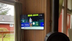 Una sala de estar con una televisión mostrando un evento deportivo, un mueble de entretenimiento de madera y una estufa de leña.