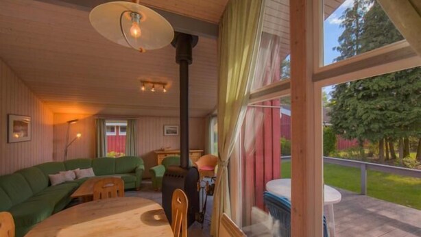 A cozy cabin interior with a wooden dining table, green sofa, and a large window overlooking a garden.