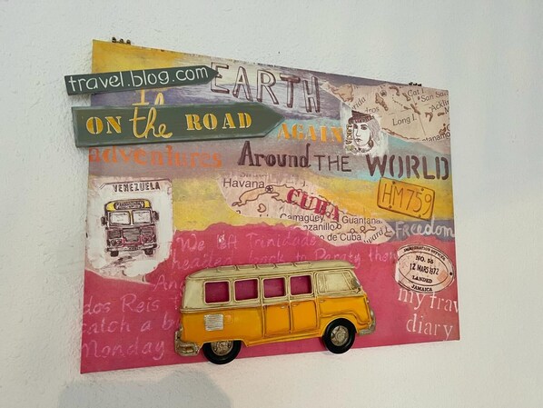 A travel-themed artwork with text and illustrations, including a bus and a vintage car, on a textured wall.