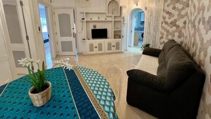 A living room with a sofa, a TV, and a patterned rug. There is a door leading to another room and a plant on the rug. - Port Cambrils 11004 (Cambrils)