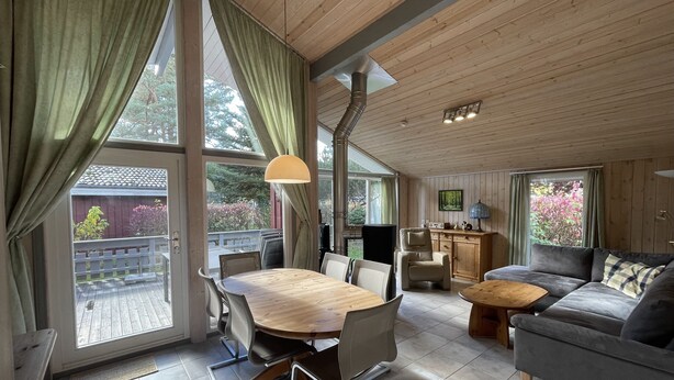 A spacious living area with a wooden dining table, comfortable seating, and large windows offering views of the outdoors.