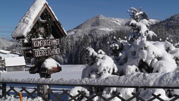 Ein Holzchalet mit einem Schild, das es als ‚Komfortzimmer Oberfeichtner‘ kennzeichnet, in einer verschneiten Berglandschaft.