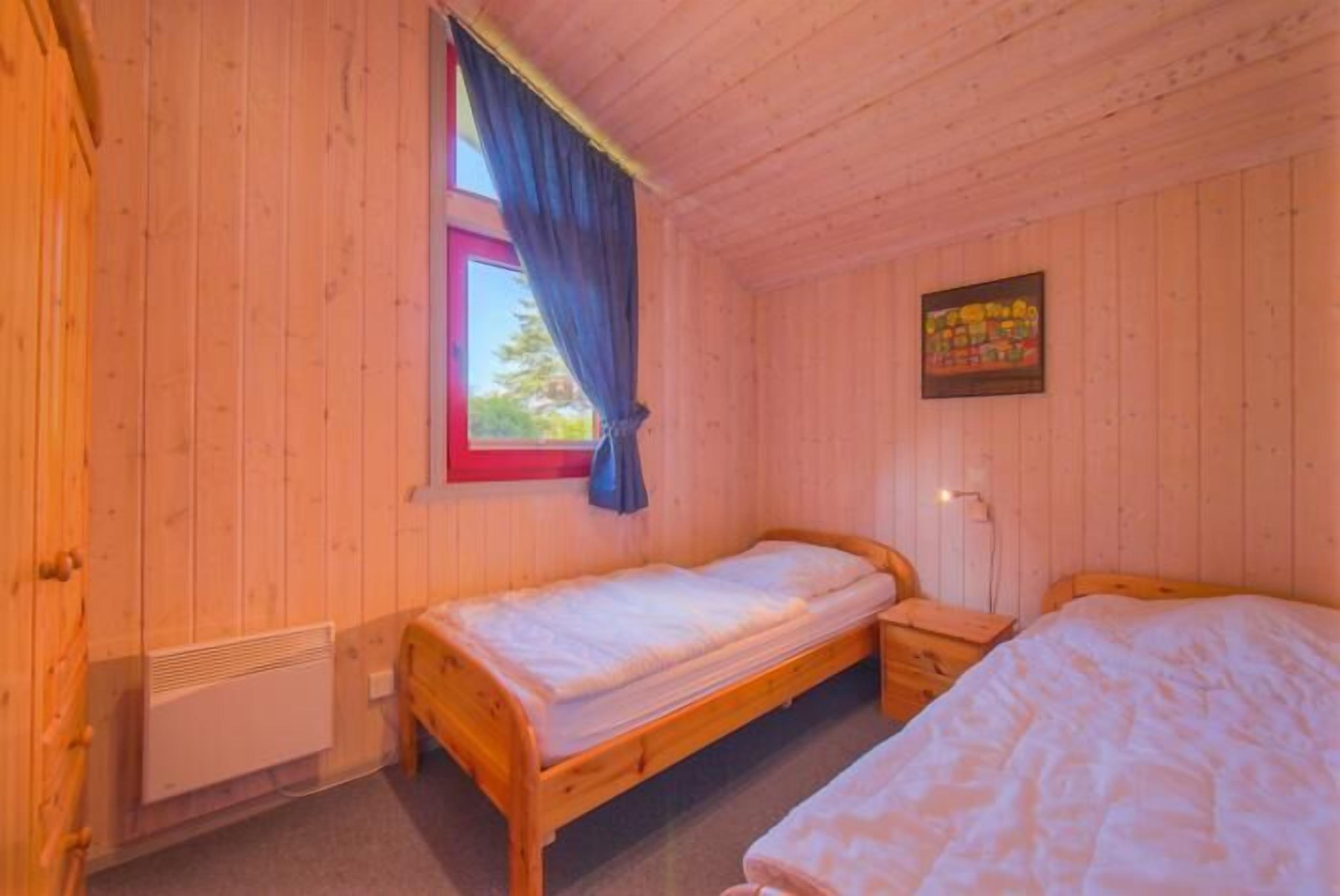 A wooden cabin room with two single beds, a window with curtains, a radiator, and a wall-mounted light fixture.