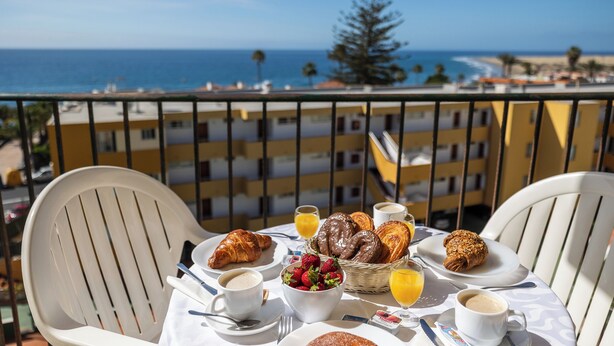 A table set with breakfast items, including croissants, fruit, and beverages, on a balcony overlooking a beach and ocean. - Apartamento Los Ficus (San Bartolomé de Tirajana)