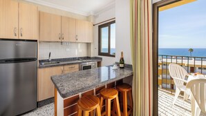 A kitchen with a stainless steel refrigerator, wooden cabinets, a granite countertop, and a bar area with stools. There is a window with a view of the ocean and a balcony with white chairs. - Apartamento Los Ficus (San Bartolomé de Tirajana)