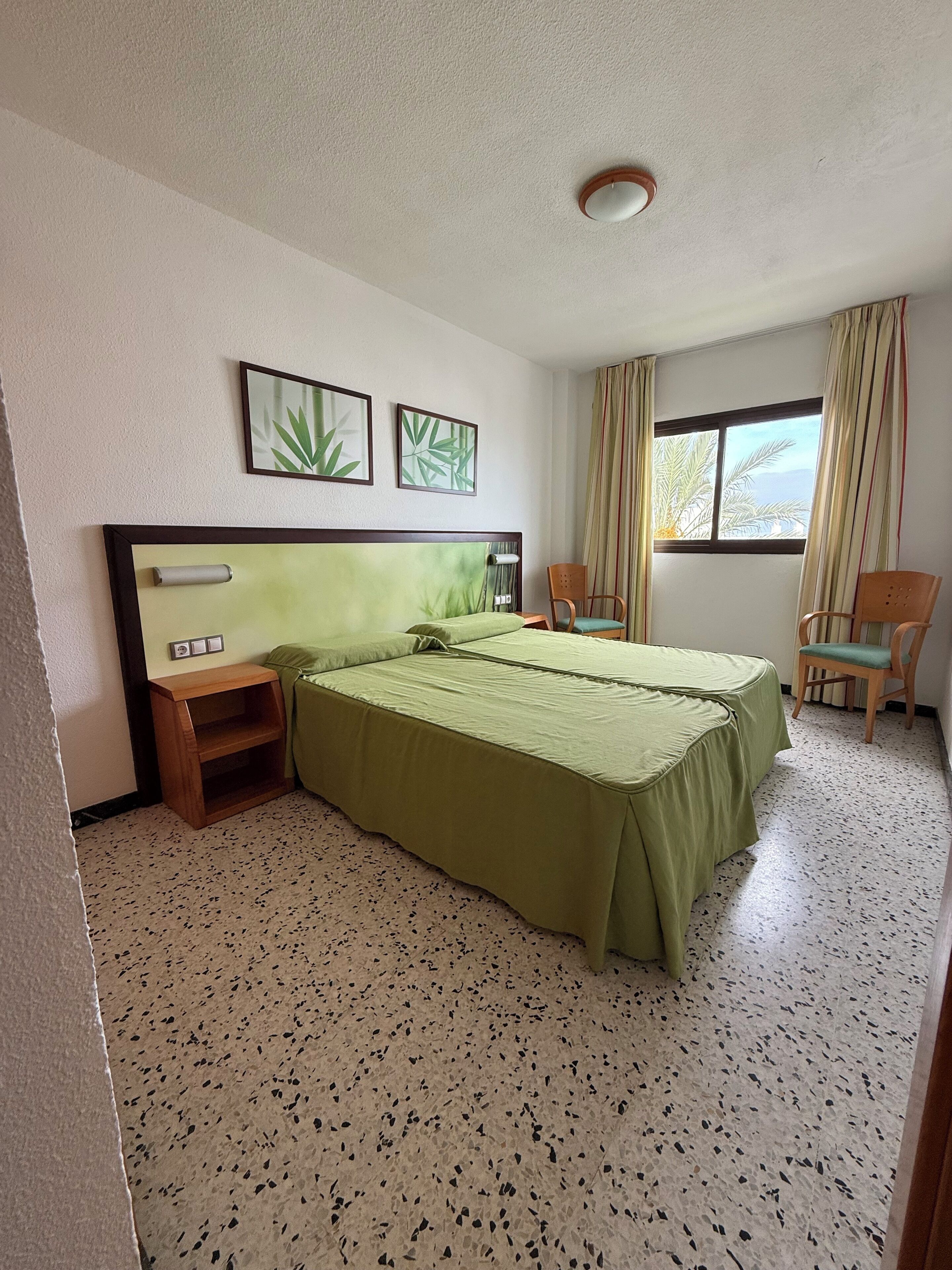 Una habitación de hotel con una cama grande, dos sillas, una mesita de noche de madera y una ventana con cortinas.