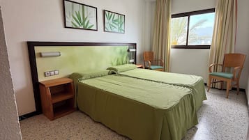 Een hotelkamer met een groot bed, twee stoelen, een klein houten nachtkastje en een raam met gordijnen.