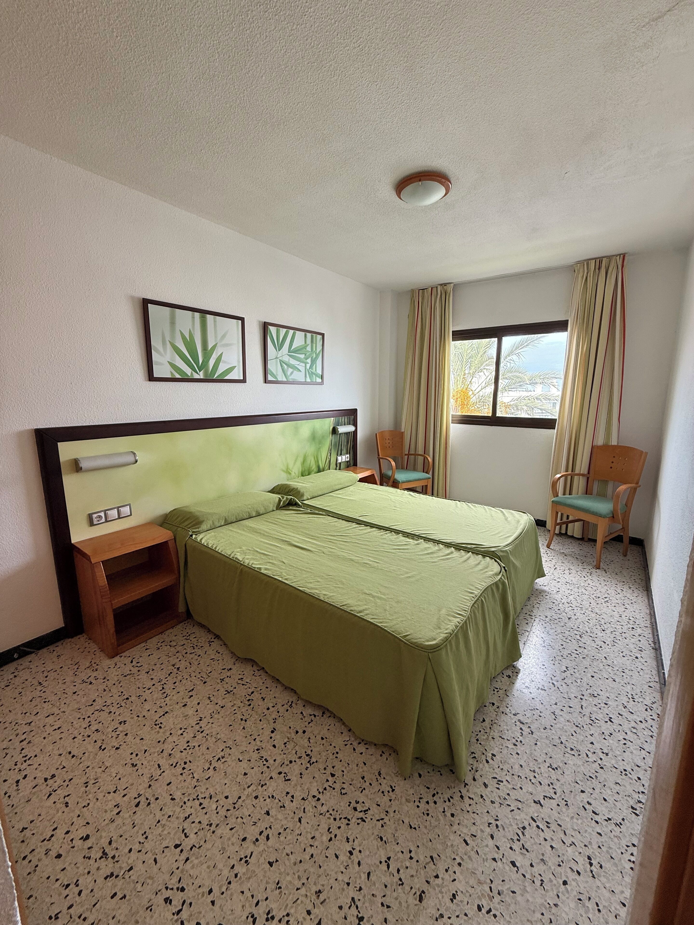 Habitación de hotel con dos camas, una mesita de noche de madera, una silla, una ventana con cortinas y dos cuadros colgados en la pared.