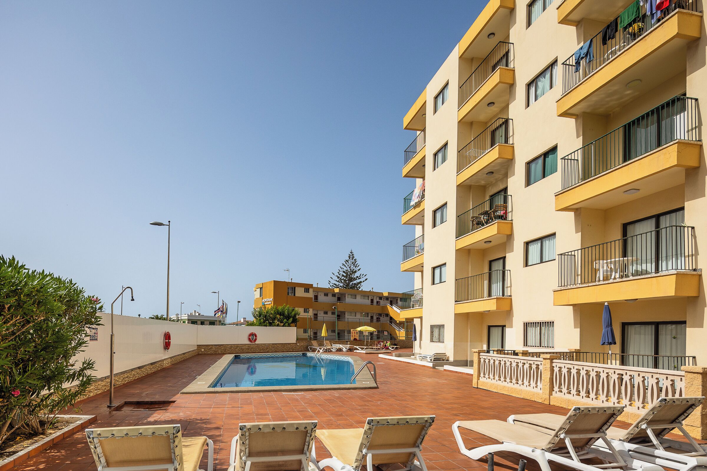 Un espace piscine avec des chaises longues, une piscine et un immeuble à plusieurs étages doté de balcons.