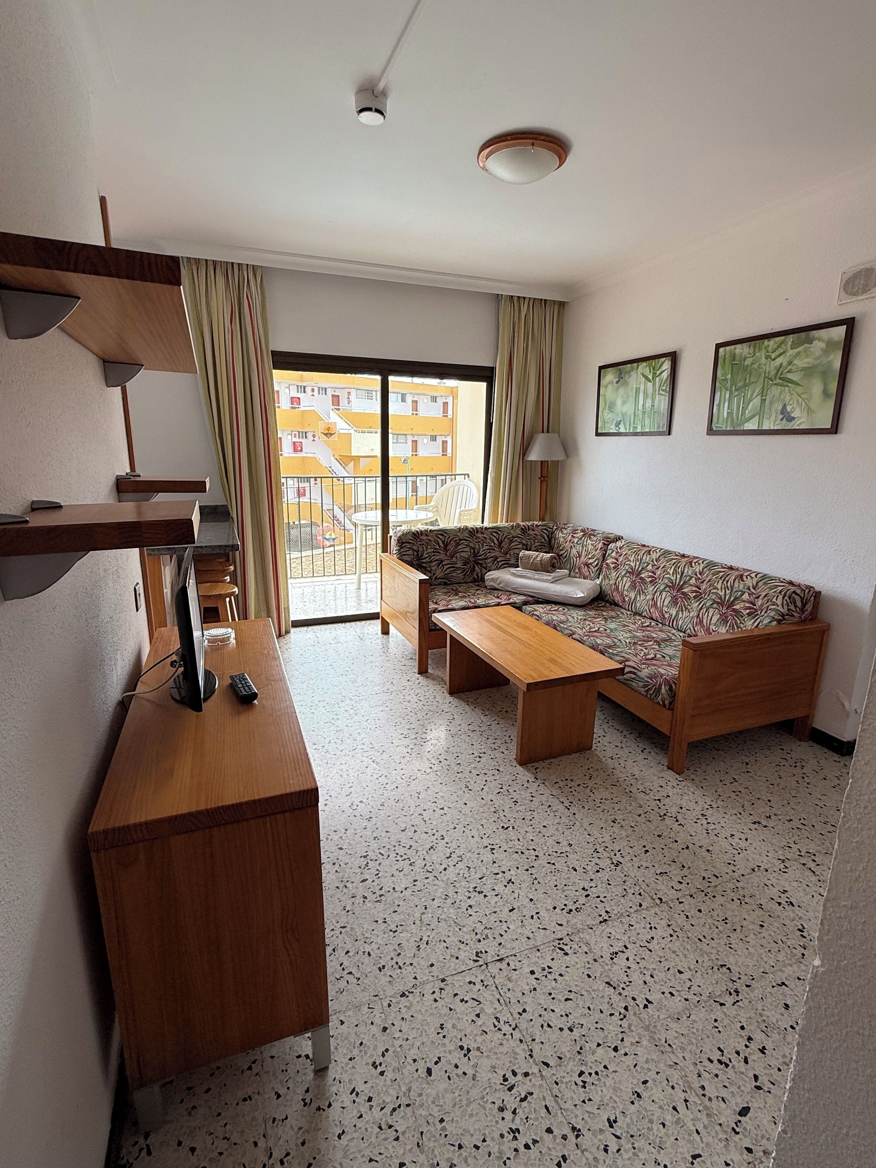 A hotel room with a sofa, a wooden desk with a computer, a small table, and a balcony with a view of buildings.