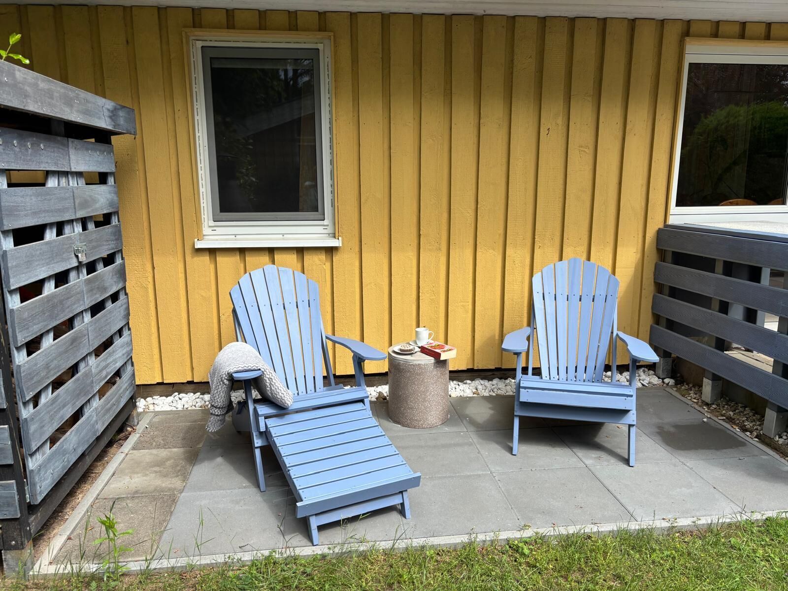 Un patio con dos sillas Adirondack, una mesita con una taza y un libro, y un cubo de basura.