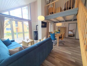 Un amplio salón con un sofá azul, piso de madera, una mesa de comedor con sillas y un entrepiso con cama y barandilla de madera.