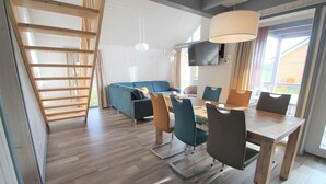 A modern living room with a wooden staircase, a blue sofa, a dining table with chairs, and a TV mounted on the wall.