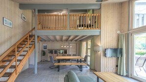 A spacious room with a wooden staircase, a dining area with a long table and chairs, a sofa, and a TV mounted on the wall.
