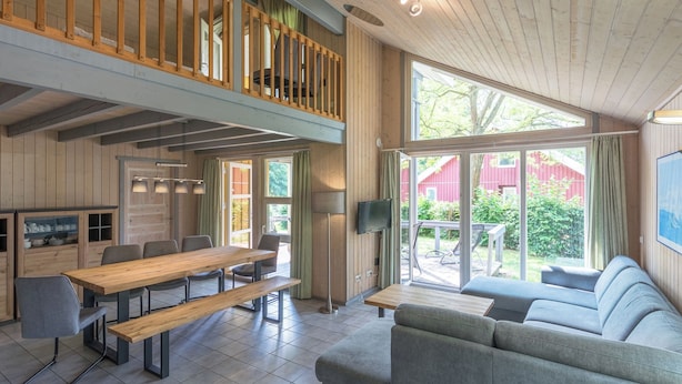 A spacious living area with a wooden dining table, benches, and a sofa. A balcony with a wooden railing is visible.