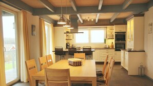 Een moderne keuken met houten kastjes, een eettafel met stoelen, hanglampen en uitzicht door de glazen deur.