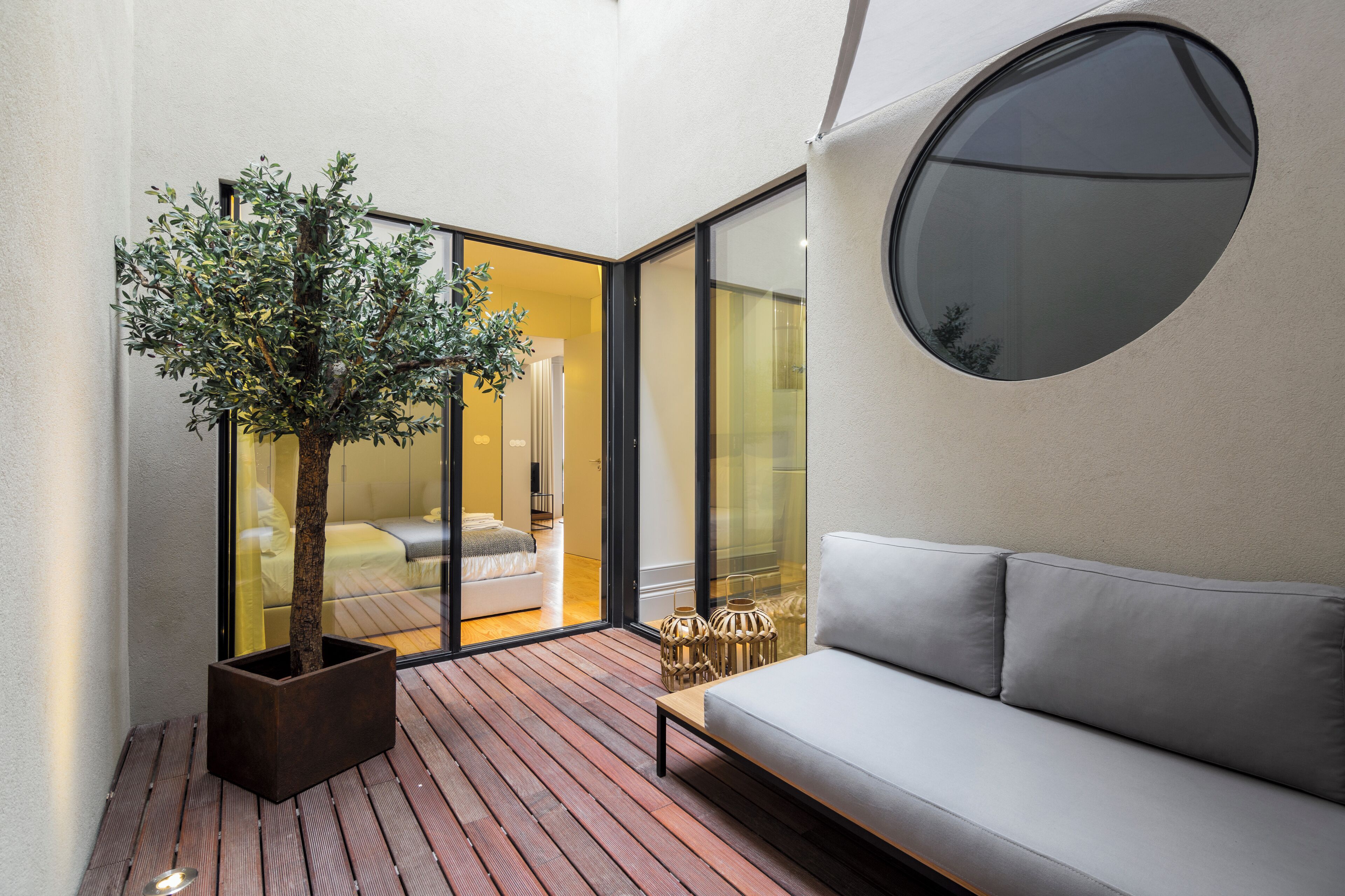A modern interior with a wooden floor, a grey sofa, a potted tree, and a large window.