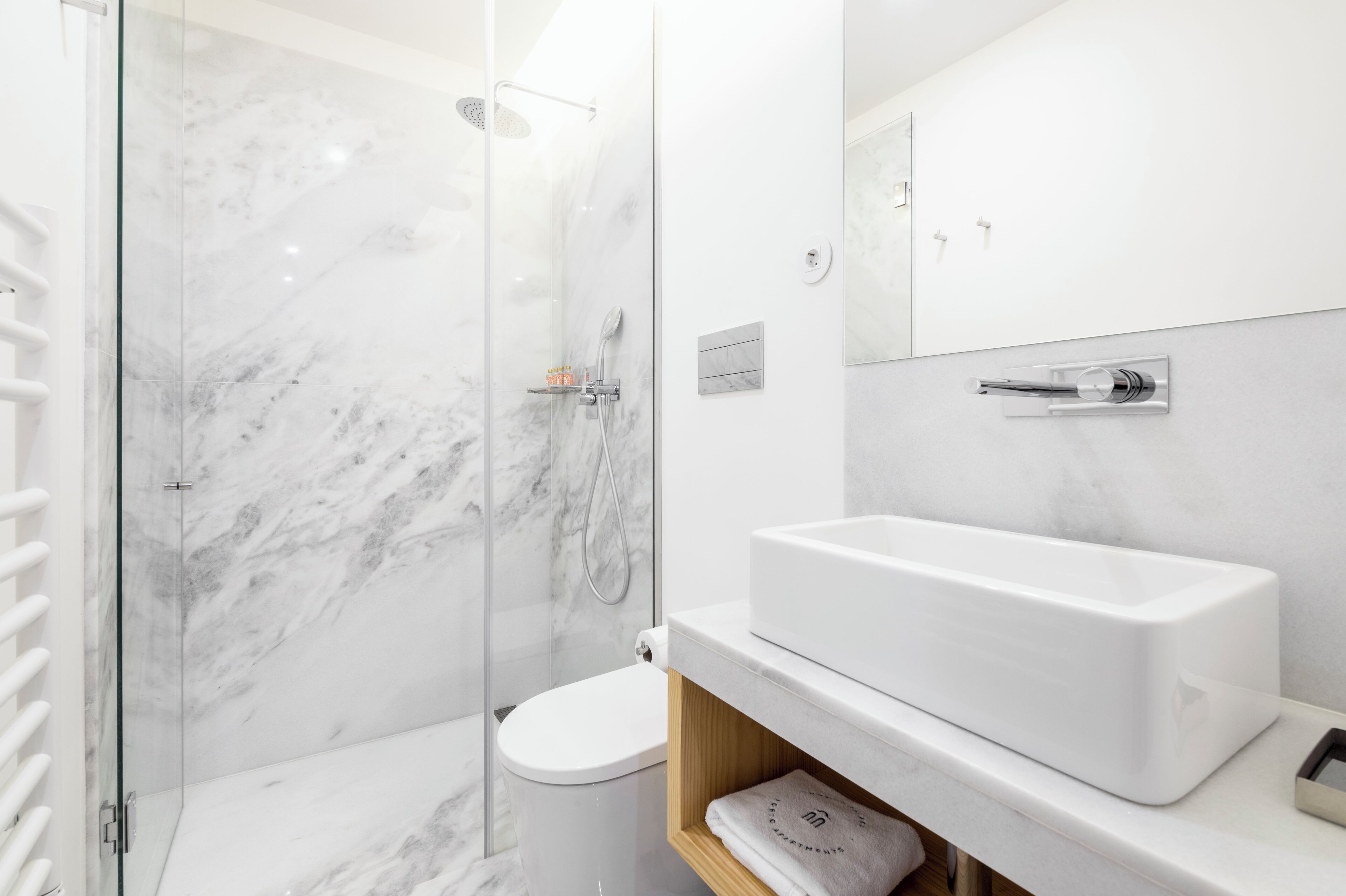 A modern bathroom with a glass-enclosed shower, a white toilet, and a large white sink with a wooden shelf below.