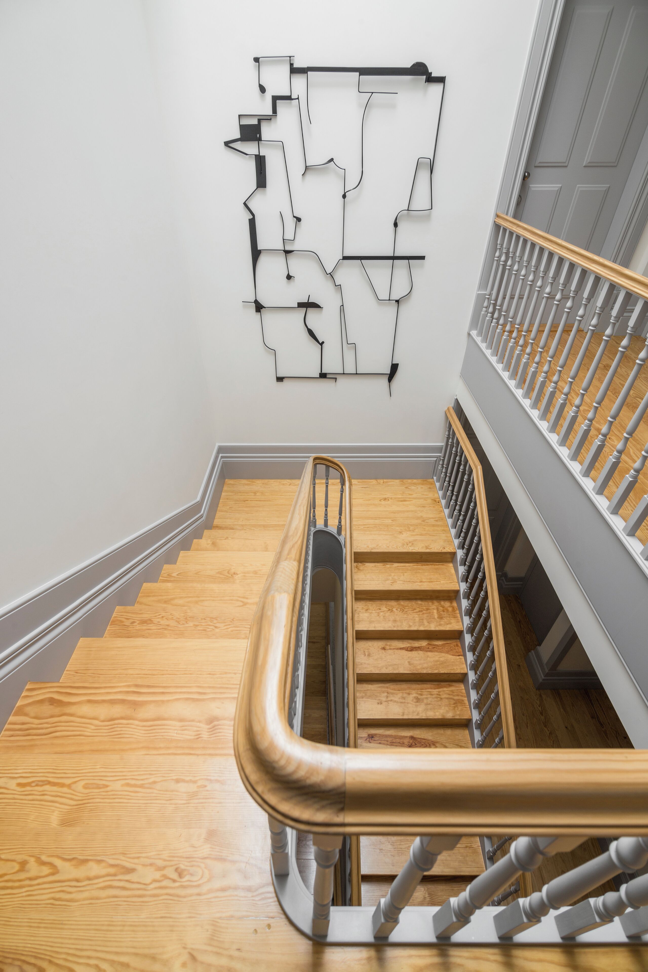 Una escalera de madera con barandilla metálica que conduce a un rellano con pared blanca y una obra de arte abstracta negra.