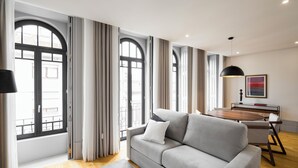 Una sala de estar moderna con un sofá gris, piso de madera y ventanas grandes con cortinas.