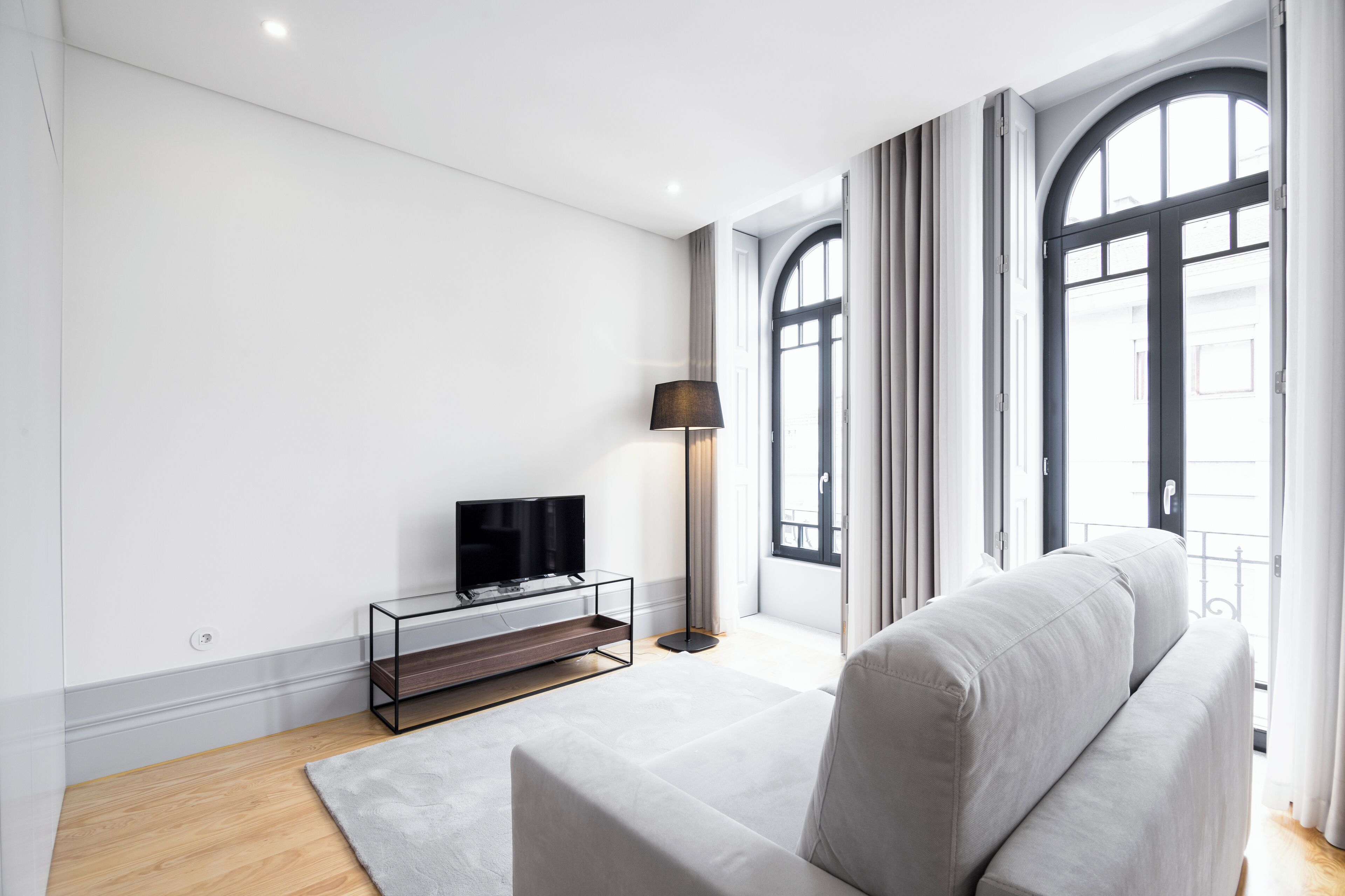 A modern living room with a grey sofa, a TV on a glass stand, and large windows with curtains.