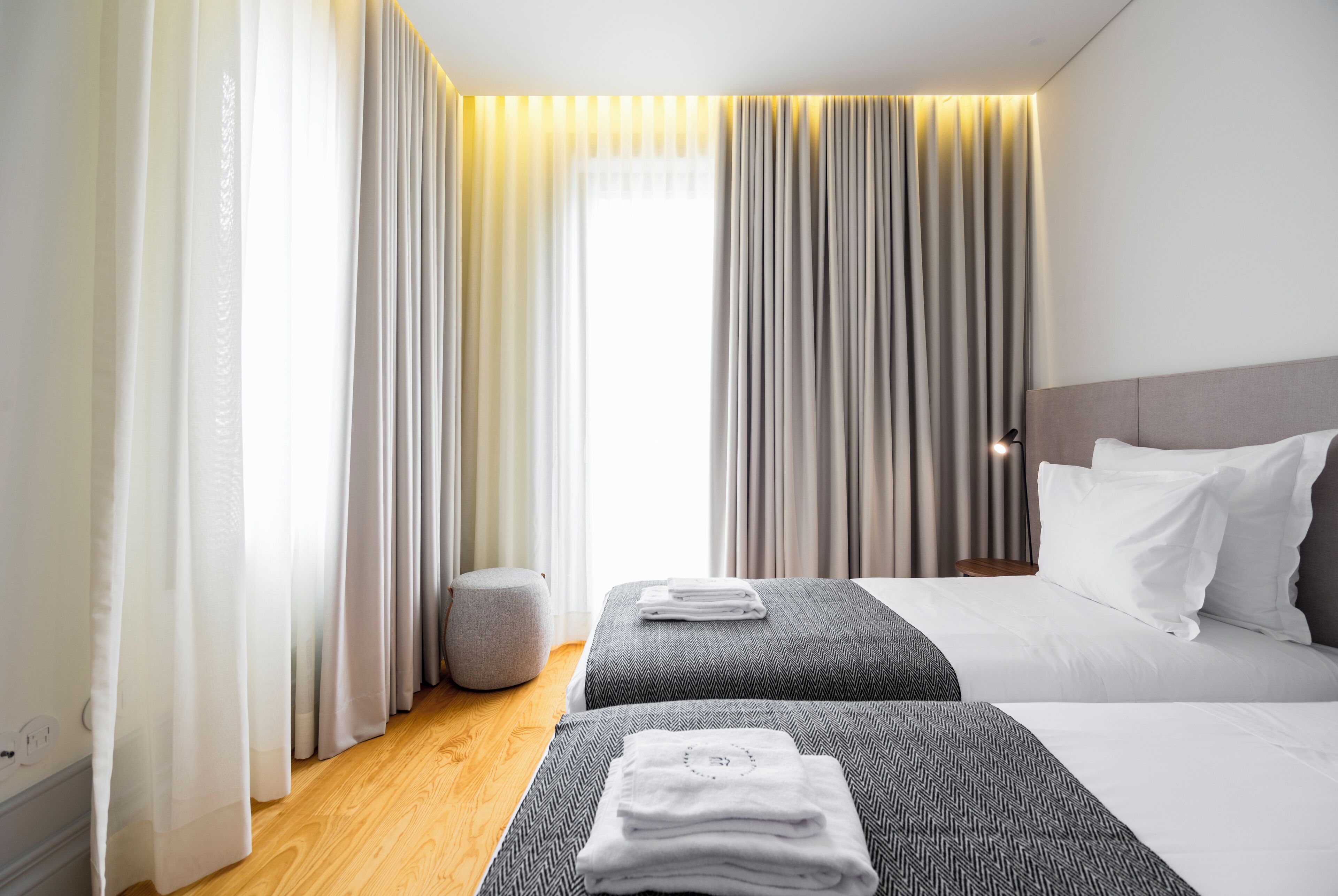 Une chambre d’hôtel avec deux lits, un pouf gris et une grande fenêtre avec des rideaux.