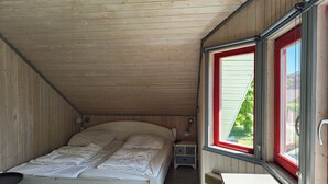 A single bed with a headboard, a bedside table, a nightstand, and a window with a red frame. - Wildgans 48 - Klassisches Skandinavisches Ferienhaus für bis zu 6 Personen (Mirow)