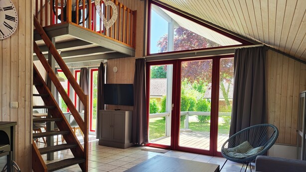 A spacious living area with a wooden staircase, a flat-screen TV, a dining table, and a large sliding glass door leading to an outdoor patio. - Wildgans 48 - Klassisches Skandinavisches Ferienhaus für bis zu 6 Personen (Mirow)