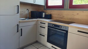 A compact kitchen with white cabinets, a stainless steel sink, a black coffee maker, a built-in oven, and a window with a view of trees. - Wildgans 48 - Klassisches Skandinavisches Ferienhaus für bis zu 6 Personen (Mirow)